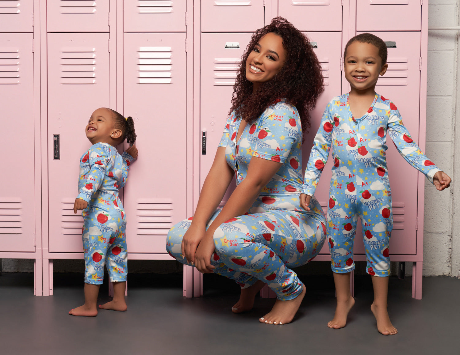 Image of a small toddler standing, with a curly haired adult woman squatting, with an older child standing on their tip-toes. The background is a line of pink lockers.